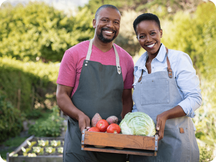 Couple all smiles in aprons holding a wooden tray with fresh farm produce outdoors